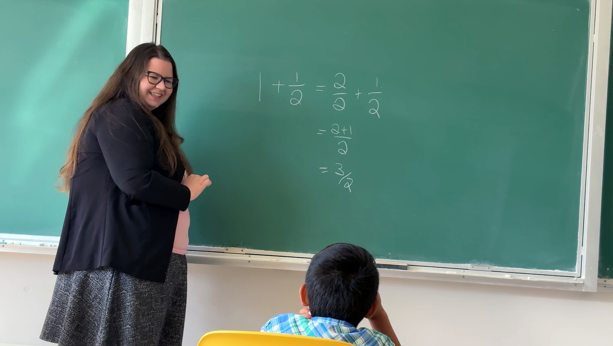 Dr. Lindsey Daniels, UBC math assistant professor of teaching stands in front of a green chalkboard with a set of simple math questions on the board. There is a kid sitting in front of her.