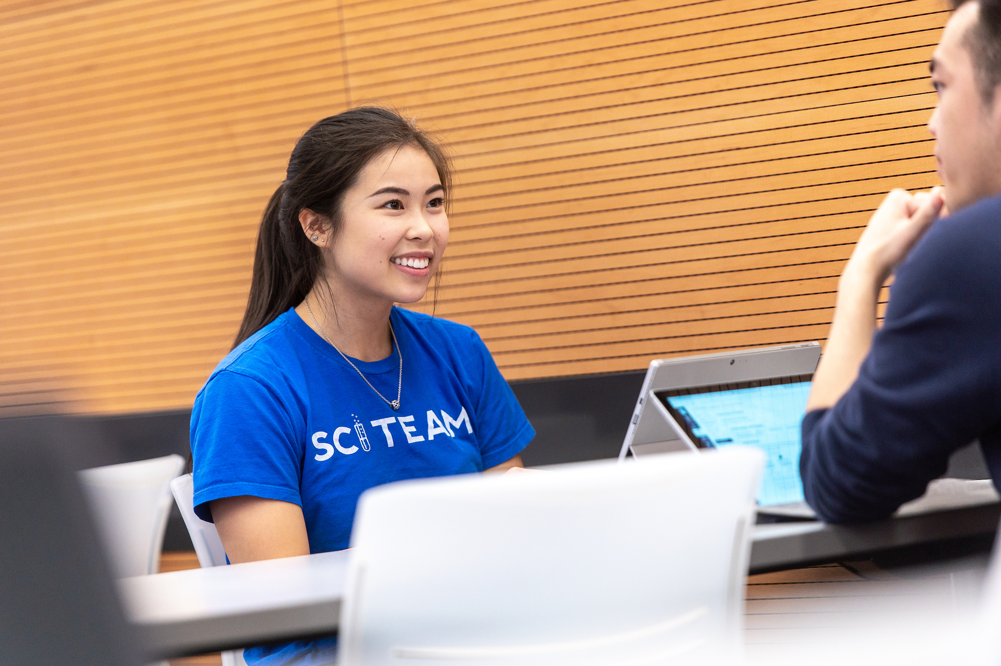 A student sits a desk wearing a blue SCI team tshirt.