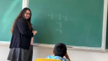 Dr. Lindsey Daniels, UBC math assistant professor of teaching stands in front of a green chalkboard with a set of simple math questions on the board. There is a kid sitting in front of her. 